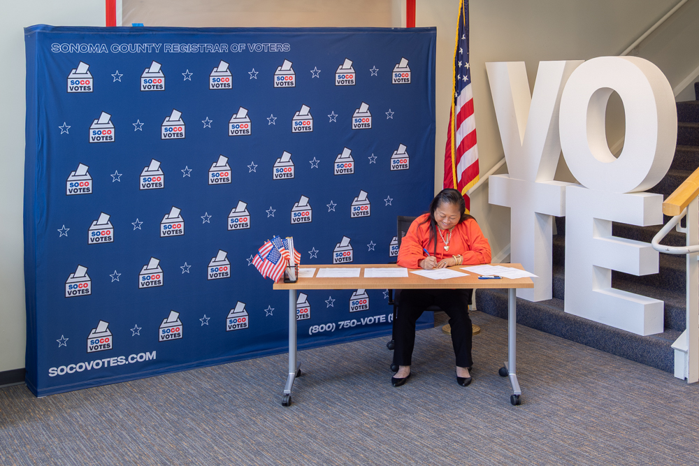 Evelyn Mendez, the Sonoma County Registrar of Votes, signs papers while sitting at a desk. In the background is a backdrop with the SoCoVotes logo repeating, an American flag, and large white letter blocks spelling out VOTE.